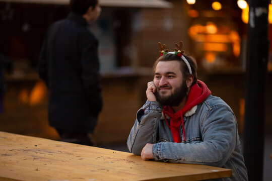 Horizontal street city photo. White man with beard, carnival deer horns in denim jacket sits at cafe table on Christmas fair, holiday market thinks about gifts for his family, wife, mother. Happy New