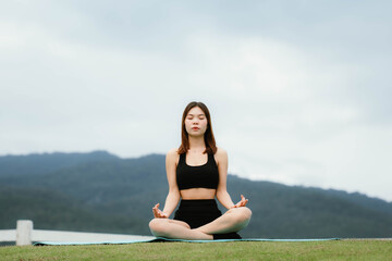 A woman meditating outdoors on a grassy field, sitting cross-legged with eyes closed, embracing...