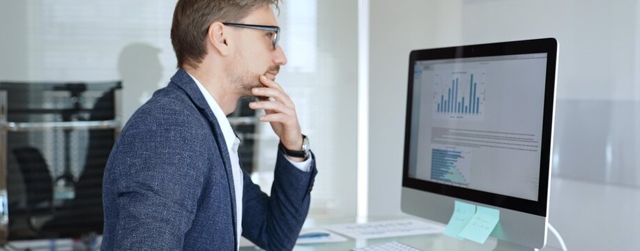Businessman in a blue jacket analyzing financial data charts on computer, making important business decisions in modern office. Business people concept - Powered by Adobe