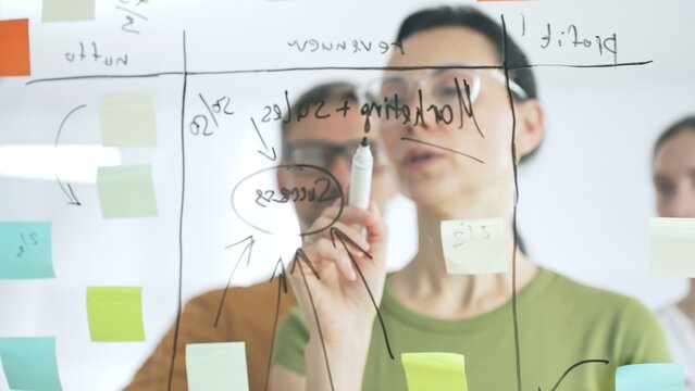 Professional team brainstorming, sharing strategy on glass whiteboard with colorful sticky notes, working collaboratively in bright office space