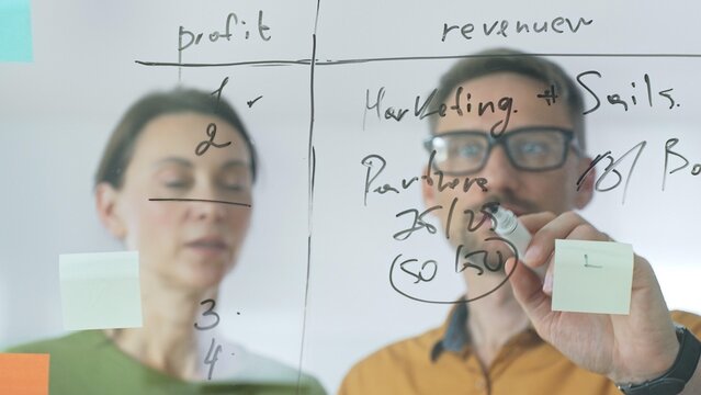 Business people brainstorming near glass board, using sticky notes for strategic planning, writing calculations, sharing innovative ideas during collaborative work session. close up