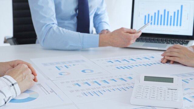 Businessman professional pointing laptop screen showing bar graphs, calculator on table while team collaborates on financial analysis and planning. Business people concept