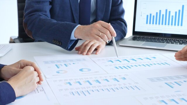 Business professional pointing at printed charts with a pen during a meeting, laptop showing bar graphs and calculator on table while team collaborates on financial analysis and planning
