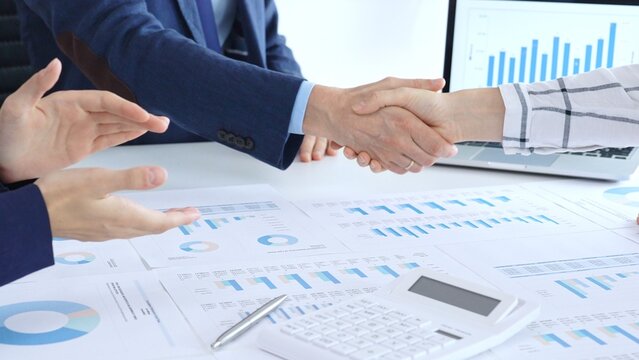 Business people shaking hands over financial documents and laptop, confirming successful partnership, agreement, or merger in a corporate office meeting - Powered by Adobe