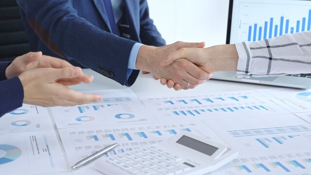 Business people shaking hands over financial documents and laptop, confirming successful partnership, agreement, or merger in a corporate office meeting