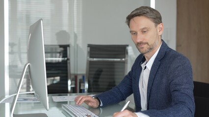 Businessman in a blue jacket working on computer, typing and writing notes in modern office, managing tasks, multitasking. Business people concept