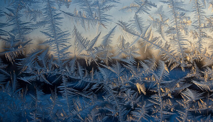 Close-up of intricate fern-like frost patterns on a window pane, illuminated by a soft, warm light from the background, creating a beautiful winter scene.