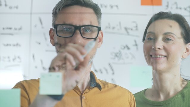 Two business people colleagues are analyzing notes on a transparent whiteboard during a brainstorming session, working together to develop innovative solutions and strategies for their deal