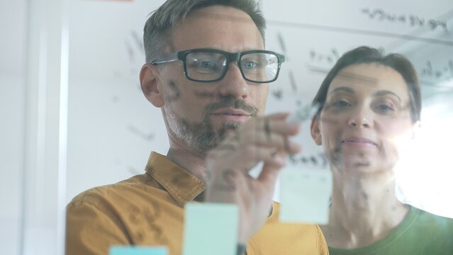 Professional team members brainstorming strategy, writing notes on transparent whiteboard with colorful sticky papers during collaborative meeting in office. Business people at work