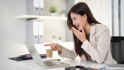 Asian businesswoman laughs pointing laptop screen enjoying online communication modern office environment professional workspace happy successful female worker tech interaction