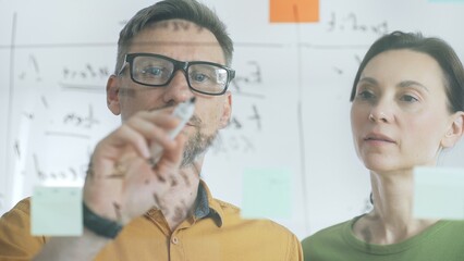 Professional businesspeople collaborating, reviewing data on transparent whiteboard, developing strategic corporate solutions in modern collaborative workspace. Business people concept