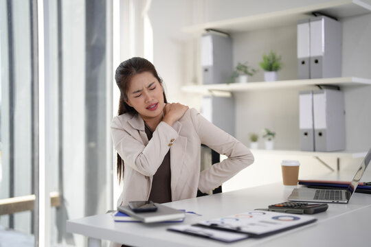Asian adult businesswoman suffers shoulder back pain working at modern office desk experiencing discomfort workplace stress health issues poor posture