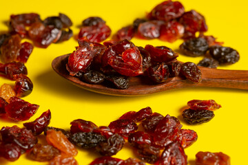 A macro close-up of a dark wooden spoon filled with a mixture of dark raisins (dried grapes) and bright red, translucent dried cranberries. The fruits are scattered on a contrasting, vibrant yellow ba
