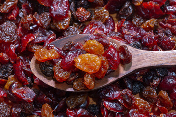 A top-down close-up shows a wooden spoon holding golden raisins, dark raisins, and dried cranberries, resting on a larger pile of the mixed dried fruit.