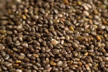 A close-up, angled shot captures a pile of dry chia seeds (Salvia hispanica). The seeds, showing...