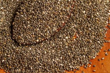 A top-down close-up shows a wooden spoon filled with dry chia seeds (Salvia hispanica), resting on a larger pile spread across a wooden surface.
