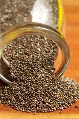 A macro, vertical close-up of chia seeds spilling out of the mouth of a clear glass jar, forming a pile on a polished reddish-brown wooden surface.