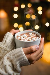 Hands holding warm cocoa with marshmallows on christmas eve with bokeh lights backgroun