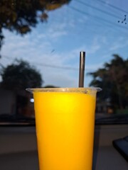 A close-up of a plastic cup filled with bright yellow mango juice, topped with a stainless steel straw, seen from inside a car against a hazy sunset sky. A refreshing drink concept.