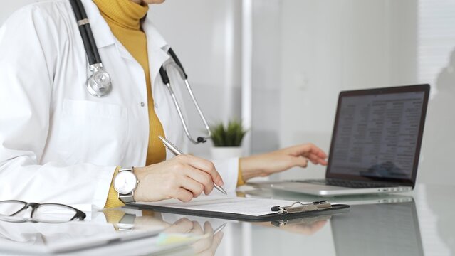 Doctor wearing stethoscope and yellow sweater under white lab coat, making notes on a clipboard in a bright medical office. Medicine, healthcare and science concept