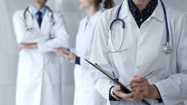A medical professional with a stethoscope writes on a clipboard while a doctor and female assistant collaborate in a clinic, discussing patient data and healthcare information