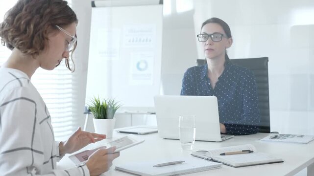 Professional women collaborating at white workspace, reviewing digital content on tablet and laptop during strategic work session - Powered by Adobe