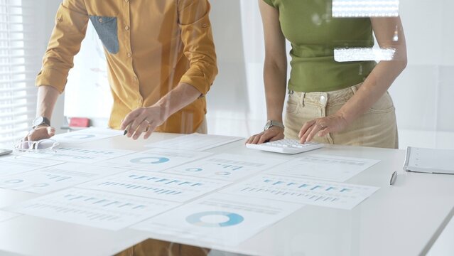 Casual business people are working together, analyzing financial data and reports using a keyboard and printed charts spread out on a table in a modern office