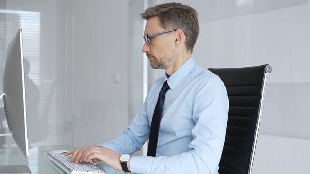 Mature professional businessman in blue shirt and tie focusing on computer screen. Brainstorming ideas and analyzing data. Working on financial strategy in modern corporate office