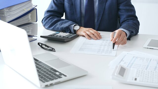 Male accountant analyzing financial documents, calculating numbers together at shared workspace with laptop and office supplies surrounding them. Audit and taxes concept