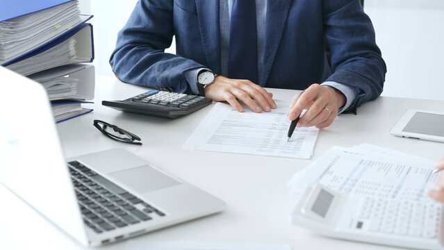 Male accountant analyzing financial documents, calculating numbers together at shared workspace with laptop and office supplies surrounding them. Audit and taxes concept