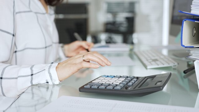 Woman calculating finances with black calculator, managing taxes, invoices, and accounting documents in office. Audit and taxes in business