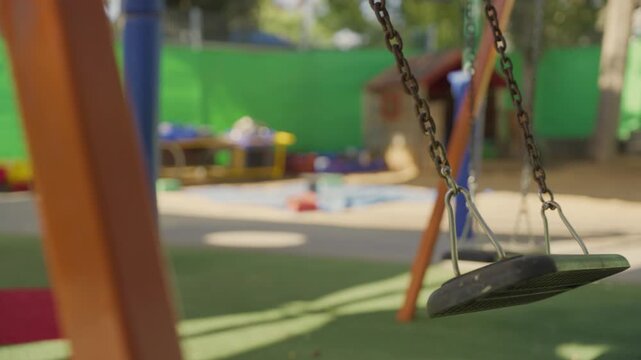 Swing moves gently in sunny playground with no children around. Empty School or Kindergarten closed, neglect, missing child, bullying aftermath, or about safety concerns , supervision, child welfare.