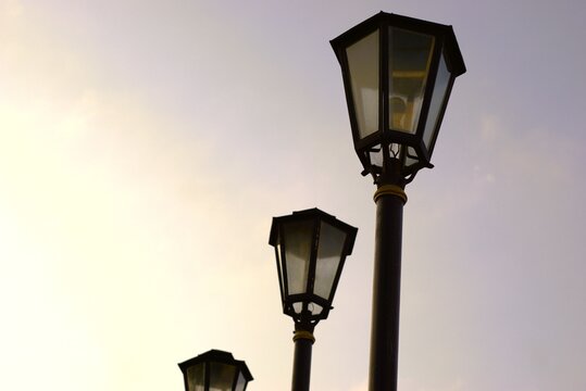 Three old-fashioned street lamps silhouetted against a soft, light-colored sky at dusk or dawn