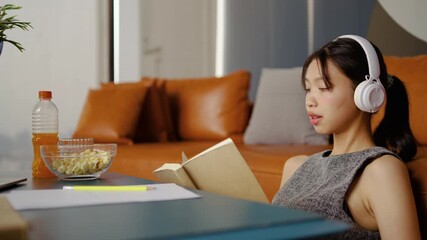 In a comfortable living room, an Asian female student concentrates on her online homework. She takes notes while listening to a teacher lecture on her laptop, while reading a book