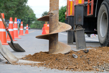 Construction workers are repairing a road by excavating and drilling the pavement to fill underground voids with specially mixed concrete. Heavy machinery and roadwork safety cones are visible.