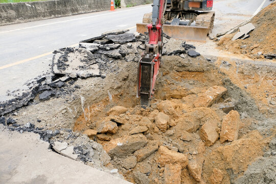 Construction workers are repairing a road by excavating and drilling the pavement to fill underground voids with specially mixed concrete. Heavy machinery and roadwork safety cones are visible.