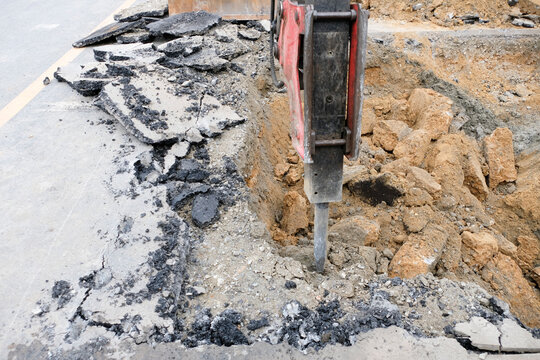 Construction workers are repairing a road by excavating and drilling the pavement to fill underground voids with specially mixed concrete. Heavy machinery and roadwork safety cones are visible.