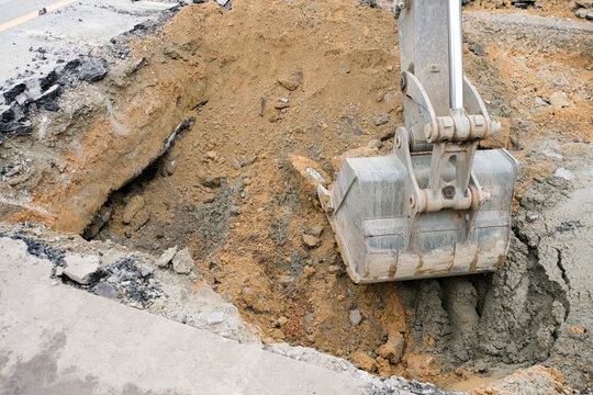 Construction workers are repairing a road by excavating and drilling the pavement to fill underground voids with specially mixed concrete. Heavy machinery and roadwork safety cones are visible.