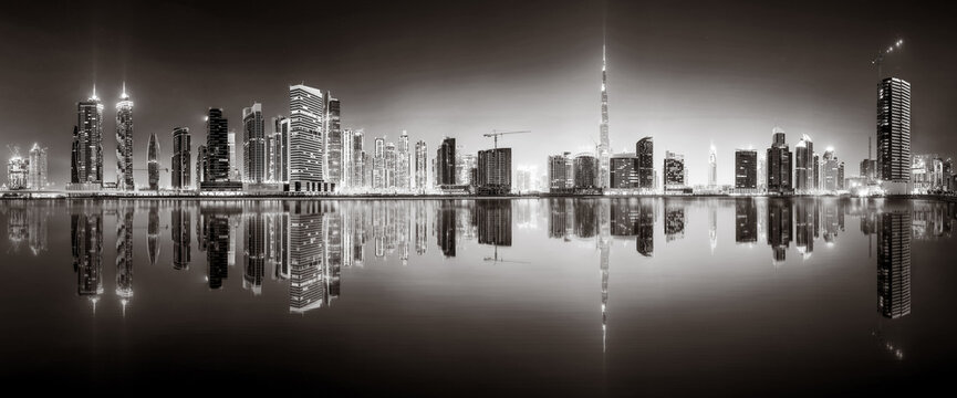 Urban city skyline with illuminated skyscrapers and waterfront railing in black and white at night, Dubai Marina bay UAE