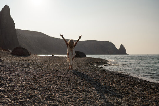 Woman, beach, freedom, joyful woman with raised arms walking along rocky seashore toward ocean during serene sunset. - Powered by Adobe