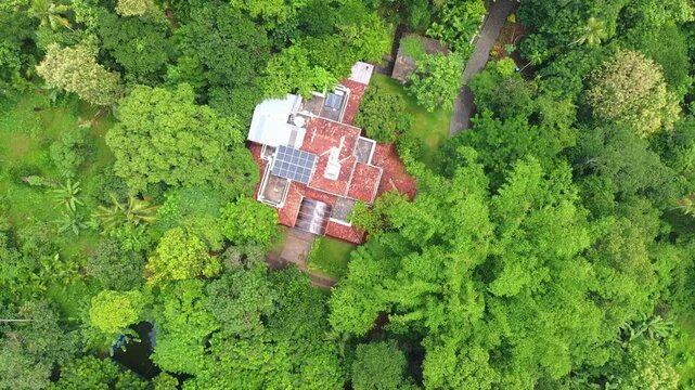 Top-down aerial view of a traditional house with solar panels, completely surrounded by dense, lush tropical rainforest or farm land in South Asia.