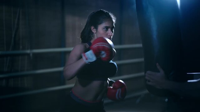 Indian young adult gen z angry fit boxer woman lady wear gloves do hand fist boxing punch beat on bag indoor dark room place modern sports female girl learn self defense martial art skill