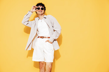 Young attractive man with short curly hair hairstyle. Smiling handsome male in casual stylish shirt and shorts clothes posing in studio, isolated on yellow. Cheerful and happy model, sunglasses