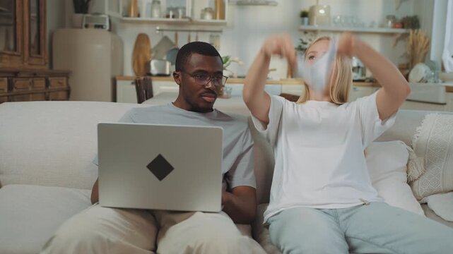 Man and woman discussing financial troubles at home, lady throwing papers in air . Portrait of young interracial spouses sitting on sofa in apartment and talking about utility bills and mortgage loan