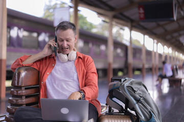 Mature man working on a laptop and talking on his smartphone while waiting with luggage on a busy train station platform, smiling and connected as he travels