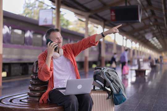 Man pointing and talking on smartphone while sitting on a bench at a train station platform with his laptop, backpack, and luggage beside him, preparing for solo travel