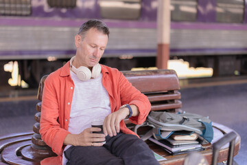 Fototapeta premium Mature man traveler sitting on a bench at a train station platform, holding a coffee cup, checking his wristwatch, and waiting for his departure, with a train in the background
