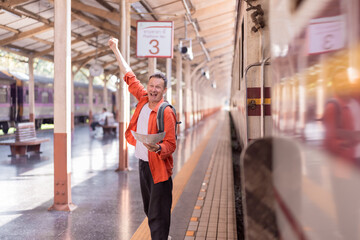 Mature man backpacker holding a map and raising his hand, celebrating success or arrival at a train station platform, feeling happy and ready for a new journey