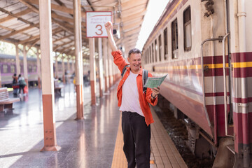 Senior man tourist happily celebrating a successful journey, holding a map and wearing a backpack, standing on a train platform with a train car beside him