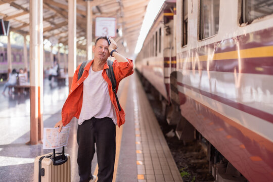 Man feeling stressed and lost while standing on a platform with luggage, looking confused and holding a map, after likely missing his train for travel